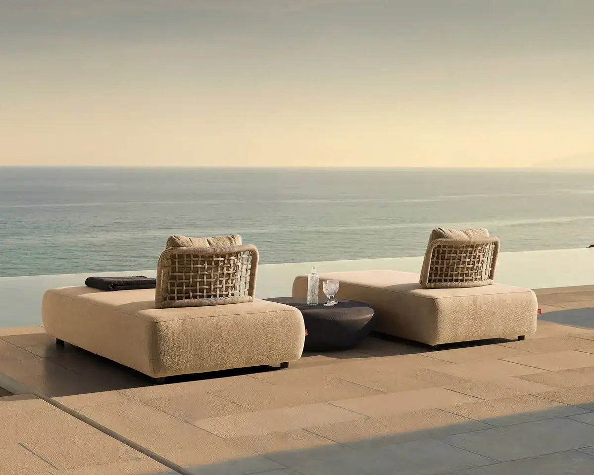 Beige woven wicker lounge chairs with cushioned seats and backrests, positioned on a stone patio overlooking the ocean.